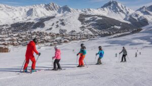 Ski instructor leading beginners on a groomed alpine slope with village and snowy mountain terrain behind them