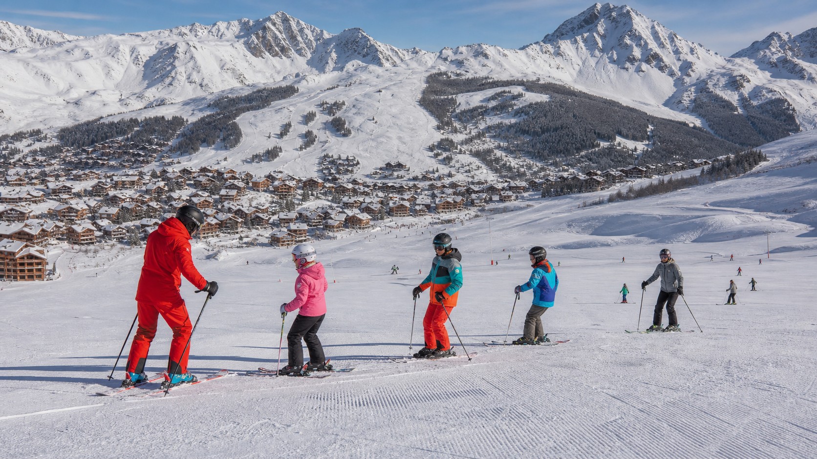 Ski instructor leading beginners on a groomed alpine slope with village and snowy mountain terrain behind them