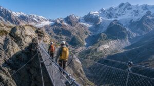 Two helmeted travelers crossing a protected alpine suspension bridge above a glacier valley